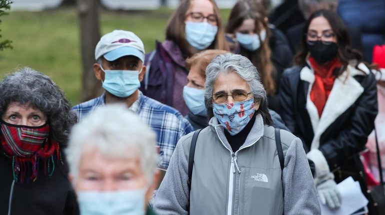 A crowd of people, most wearing masks, walk down an...